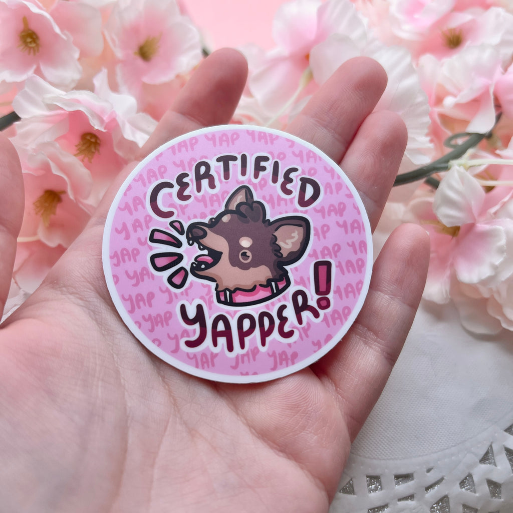 A sticker of a brown chihuahua with its mouth open, text that says "certified yapper" around it, and little yaps in the background