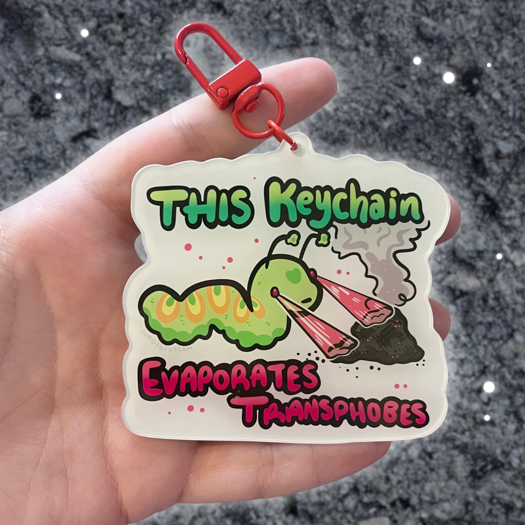 A keychain of a green worm shooting lasers out of its eyes onto a pile of ash with text that says "this keychain evaporates transphobes"