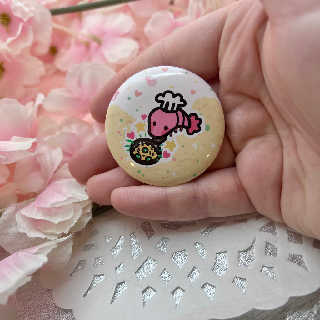 A button of a shrimp with a chef hat, holding a skillet with rice, with rice, peas, etc. in the background