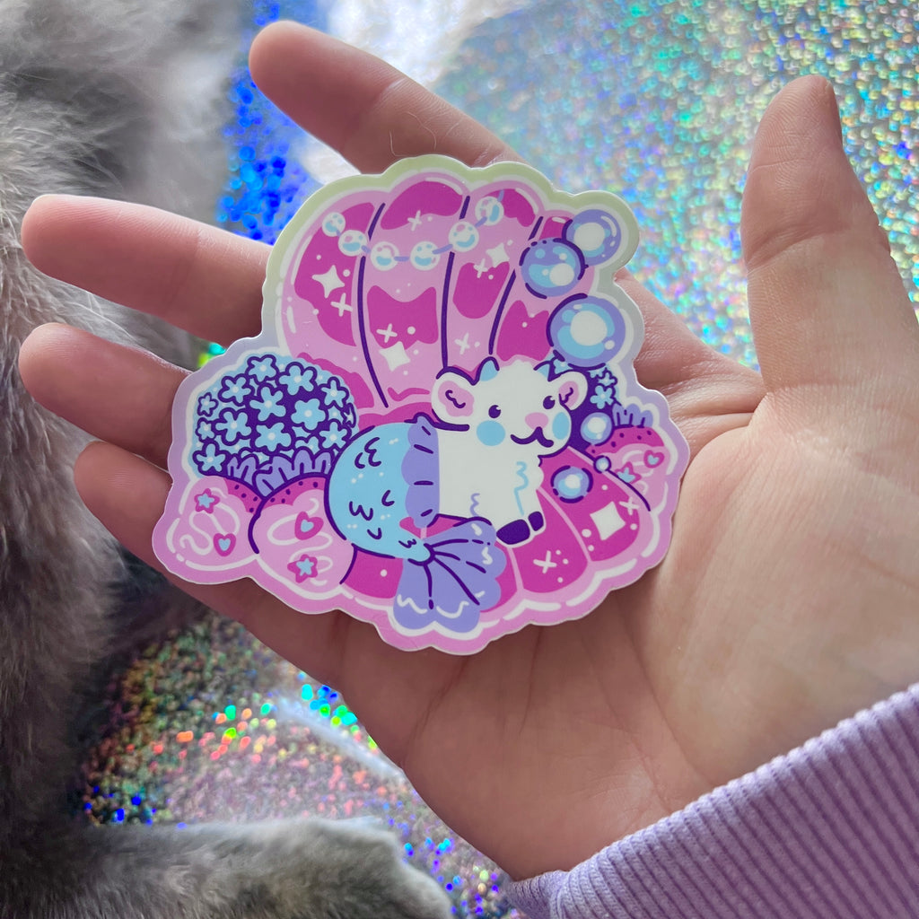 Hand holding a colorful Colorful sticker of a mermaid goat in a pearl shell  against a glittery background