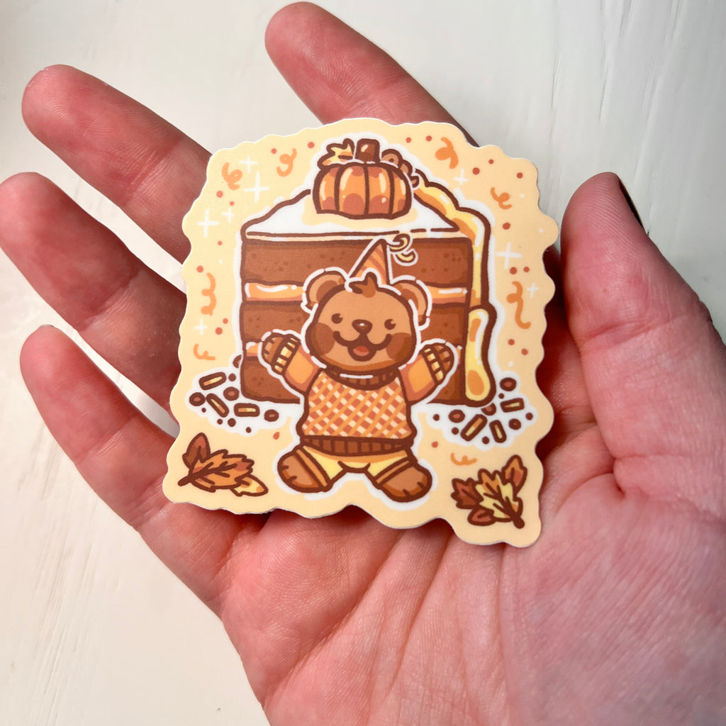 Sticker of a bear in an orange outfit with a pumpkin on top of a cake, held in a hand.