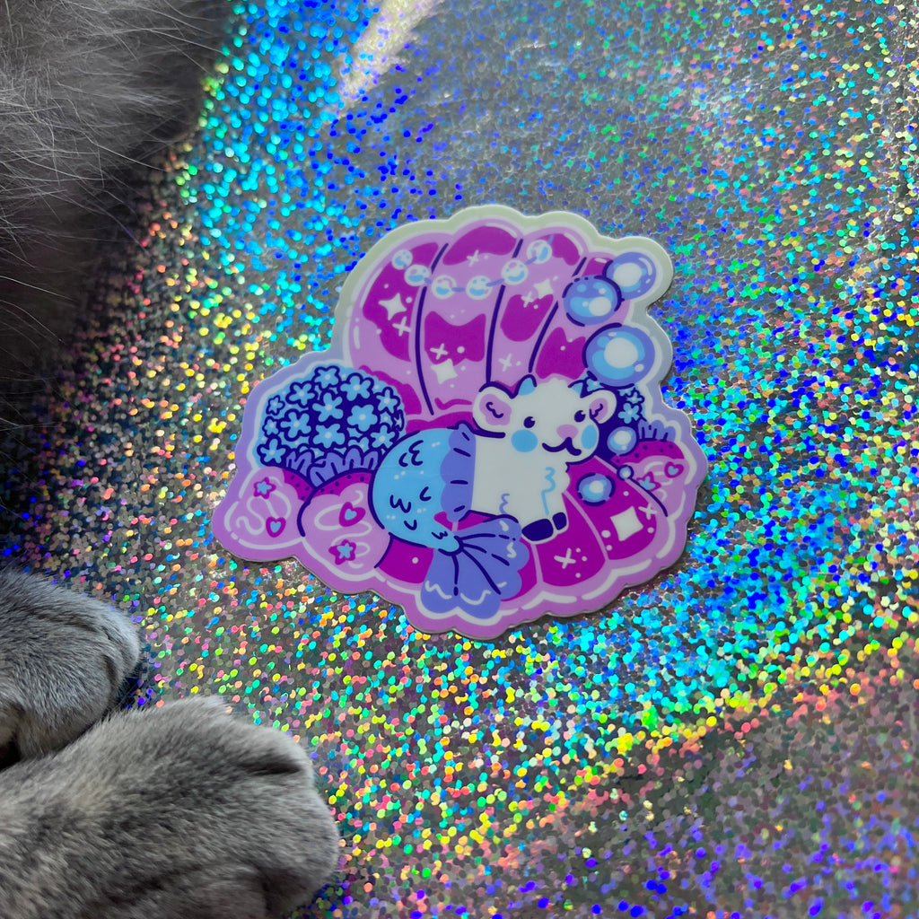 A colorful sticker of a mermaid goat in a pearl shell  on a glittery background with a cat's paw in the foreground.