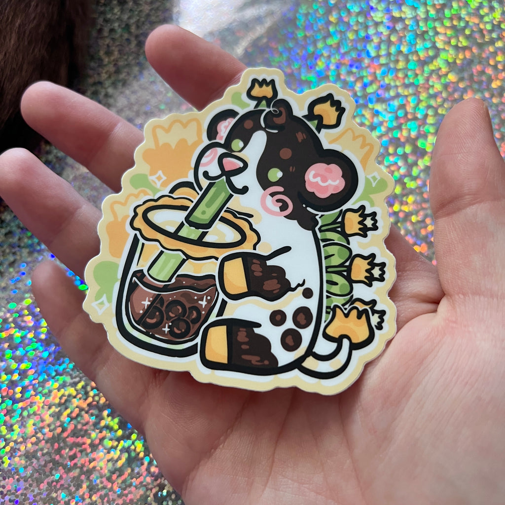 Hand holding a cartoon cow drinking boba tea sticker on a glittery background