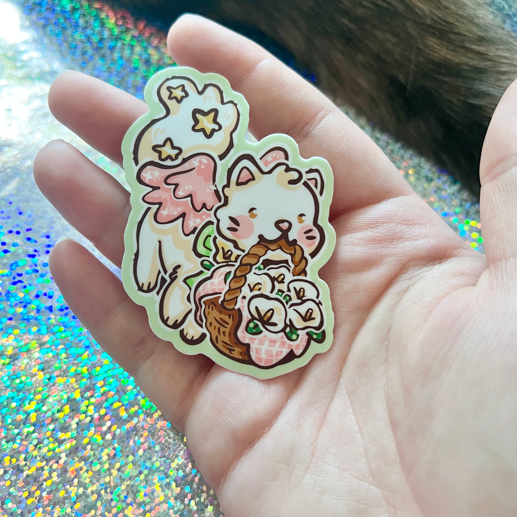 Sticker of a cat with angel wings and a basket held in a hand against a glittery background