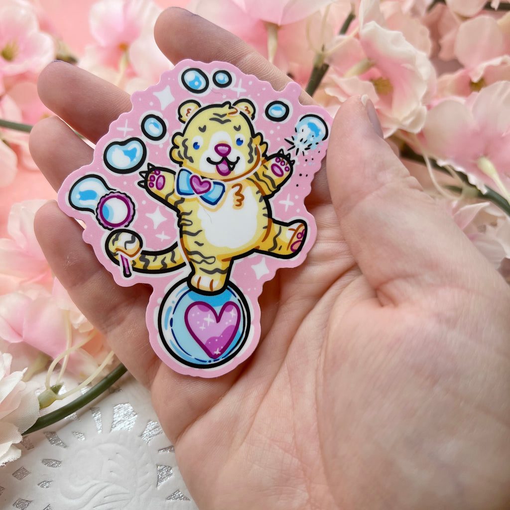 A sticker of a cute tiger balancing on a ball, bubble wand in tail, reaching up and poking the bubbles with his claws. Pink aesthetic overall.