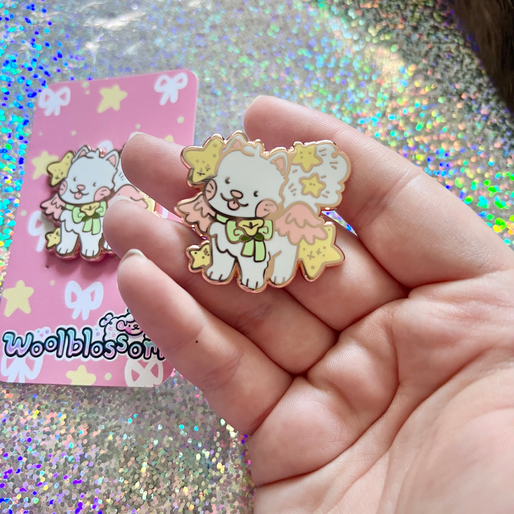 An enamel pin of a white cat with pink wings and yellow stars held in a hand with a glittery background