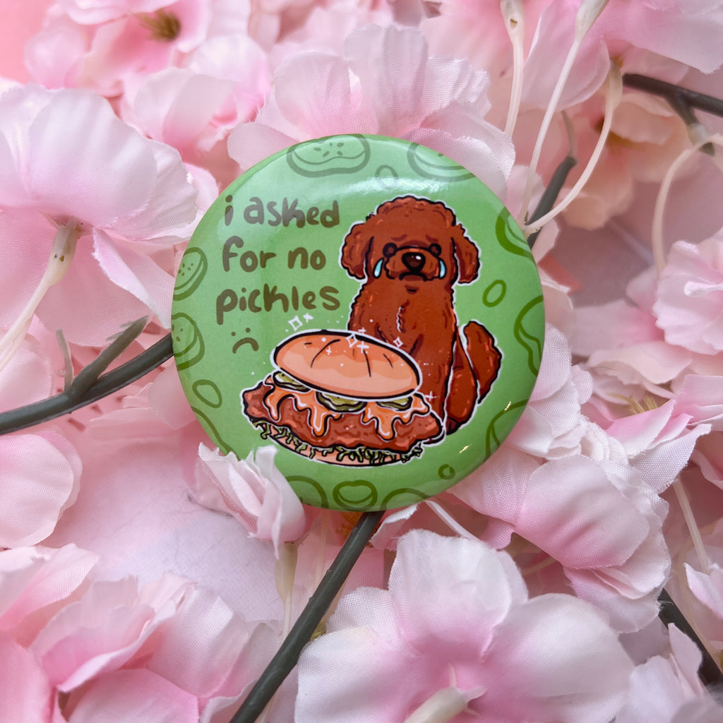 A button of a brown poodle dog sad and crying in front of a chicken sandwich that has pickles. Text says "i asked for no pickles :(" against a green background with pickle outlines