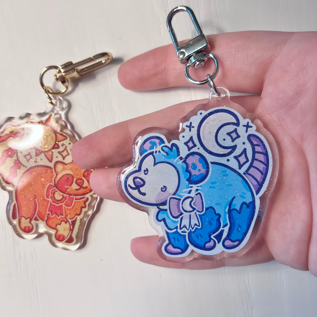 Two colorful keychains featuring cartoon sun raccoon and moon opossum held in a hand against a white background.