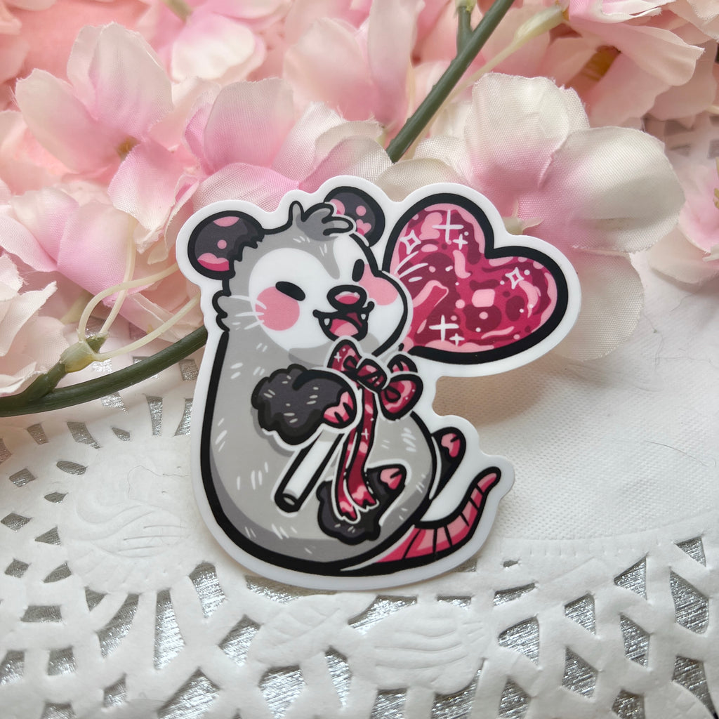 A sticker of an opossum happily holding a red heart lollipop