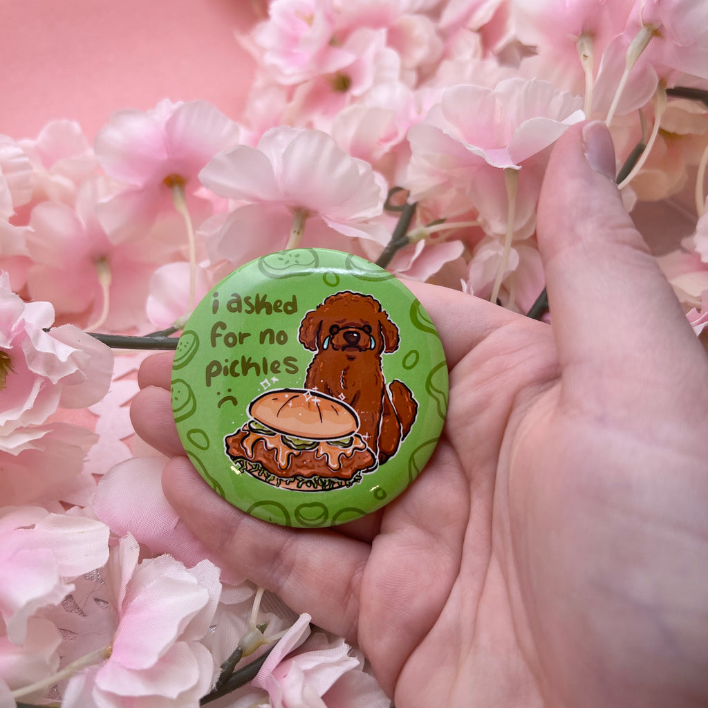 A button of a brown poodle dog sad and crying in front of a chicken sandwich that has pickles. Text says "i asked for no pickles :(" against a green background with pickle outlines