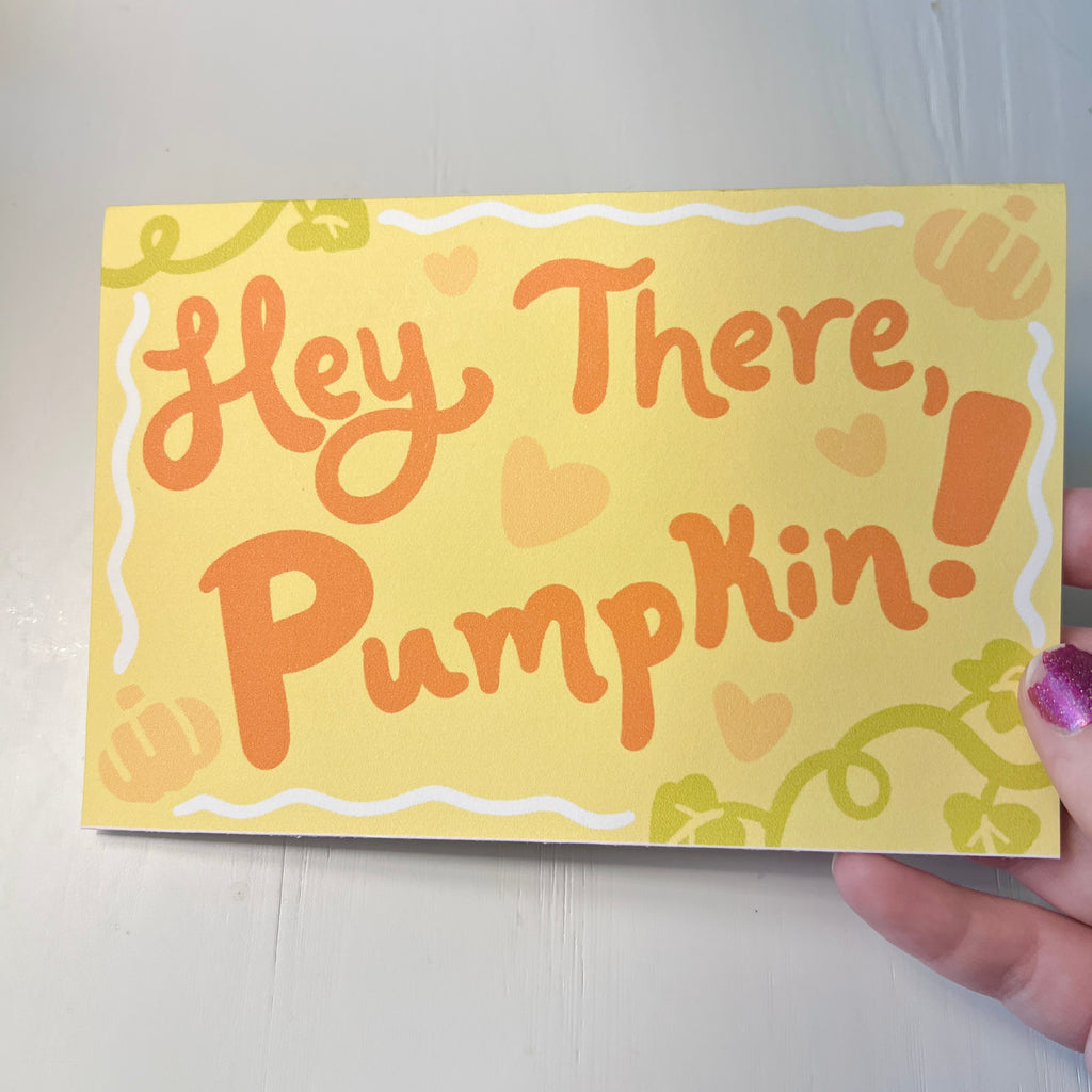 Hand holding a card with 'Hey There, Pumpkin!' text on a plain background