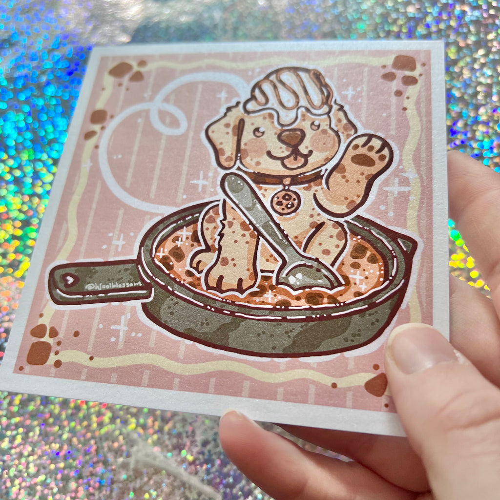 print of a puppy in a cookie skillet, held by a hand against a glittery background.