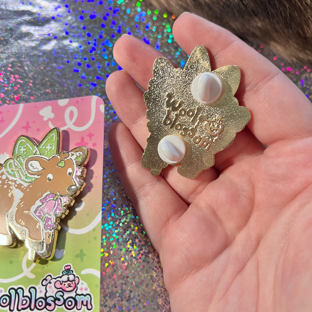 An enamel pin of a fairy deer holding a heather flower held by a hand with a colorful glittery background.