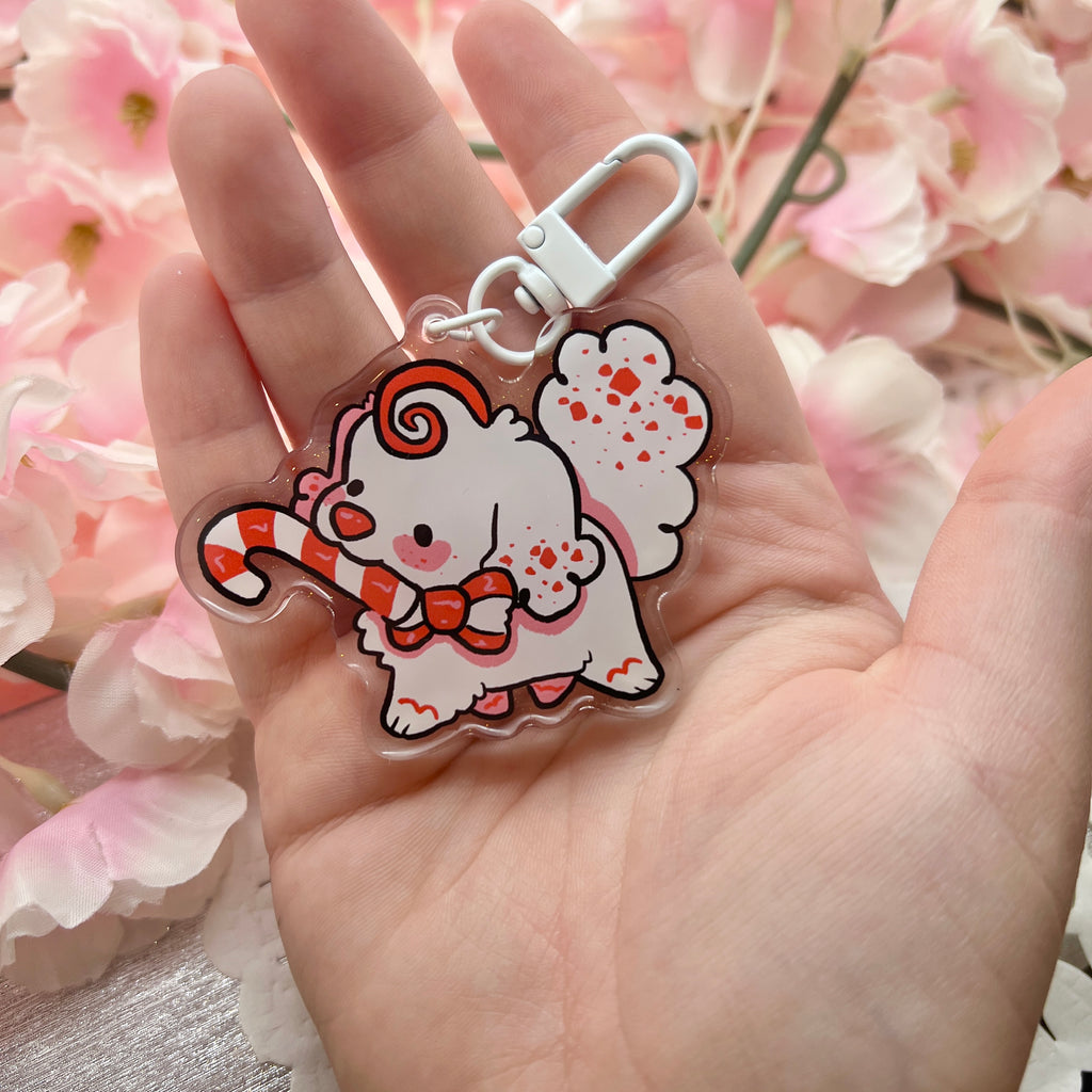 A keychain charm of a white peppermint bark themed puppy holding a candy cane in her mouth