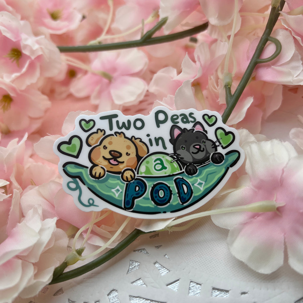 A sticker of a pea pod with a golden retriever and a black cat sticking out and text that says “two peas in a pod”