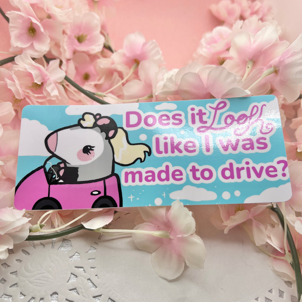 A bumper sticker of a blonde feminine opossum driving with text that says "does it look like I was made to drive"