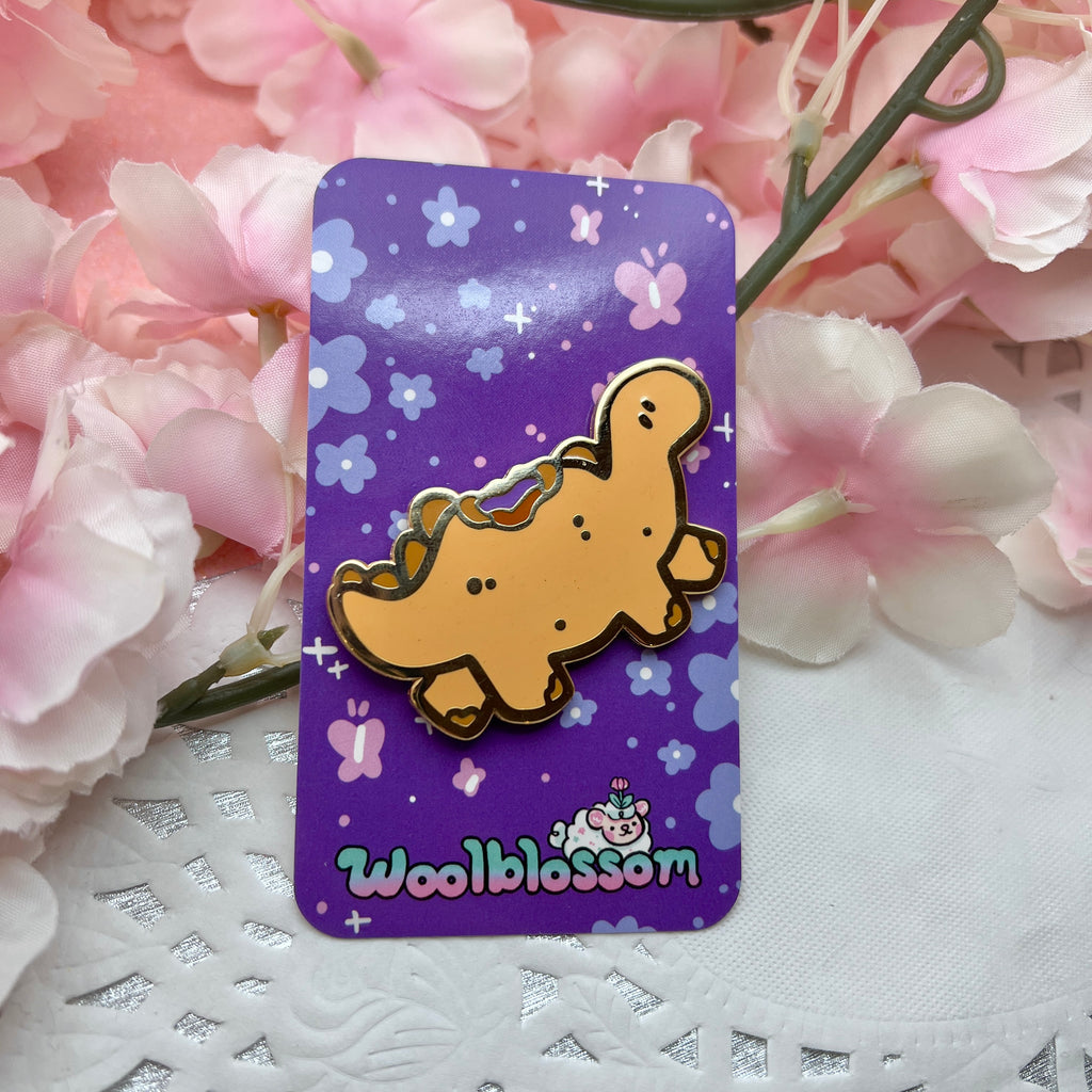 A gold enamel pin of a tan colored stegosaurus inspired dino that resembles a PBJ sandwich, the crustless kind, with a bite taken out of its back so you see the PB and jelly