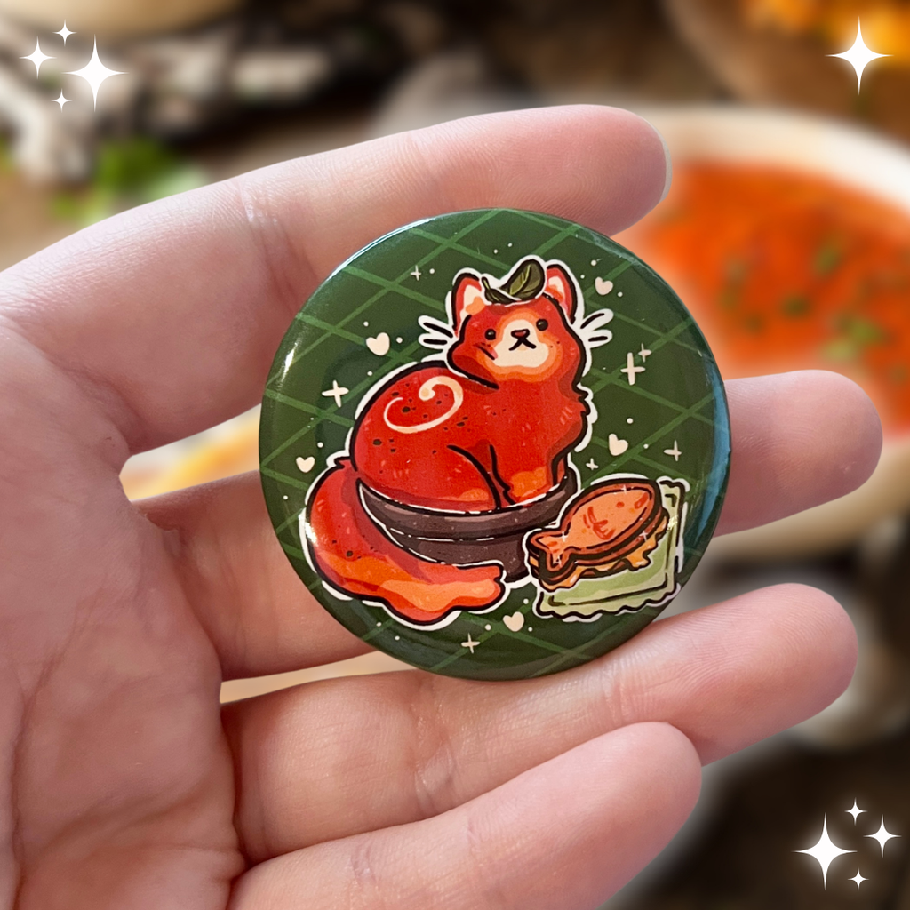 A button of a red tomato soup themed cat in a bowl, basil leaf on head, with a fish grilled cheese next to them.