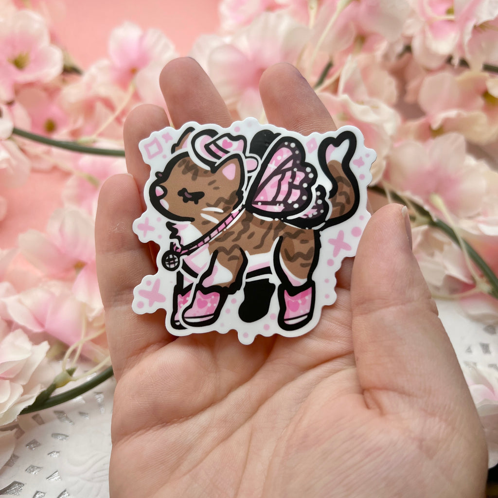 a sticker of a brown and white tabby cat with a white cowgirl hat, pink boots, and light pink butterfly wings.