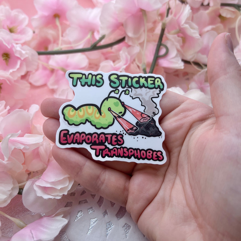 A sticker of a green worm shooting lasers out of its eyes onto a pile of ash with text that says "this sticker evaporates transphobes"