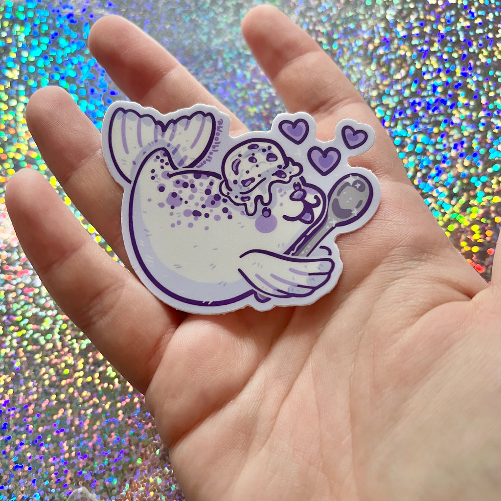 Hand holding a sticker of a cookies and cream seal against a glittery background