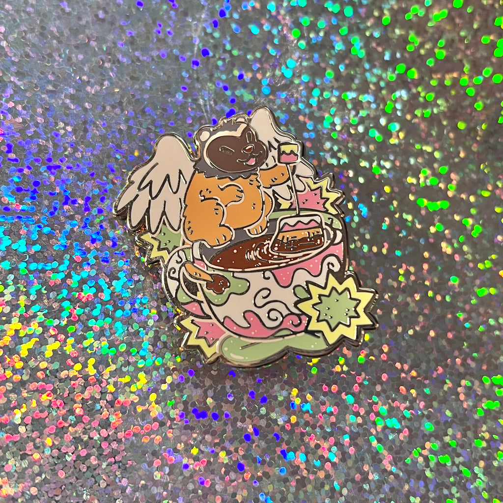 An enamel pin of a badger gryffin next to a teacup on a glittery background