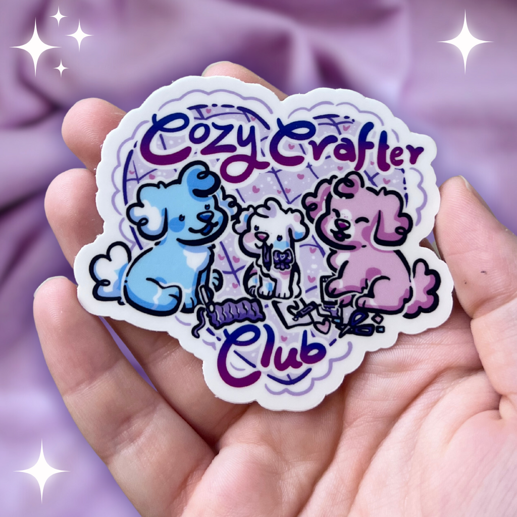 a sticker of three puppies who are knitting, doodling, and scrapbooking. One puppy is blue, one pink, and one white. There is text that says "cozy crafter club".