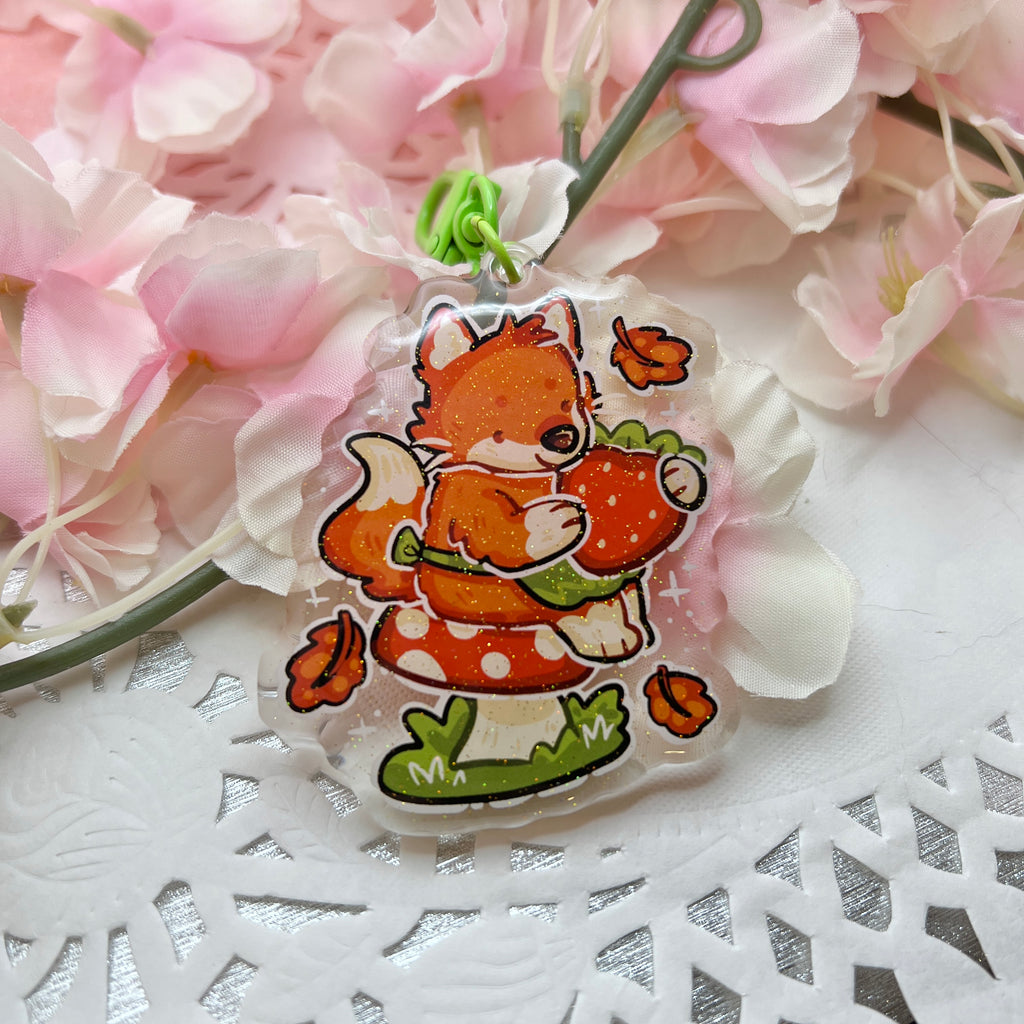 A keychain of a red fox with a little green apron sitting on a red mushroom, holding a strawberry with autumn leaves falling