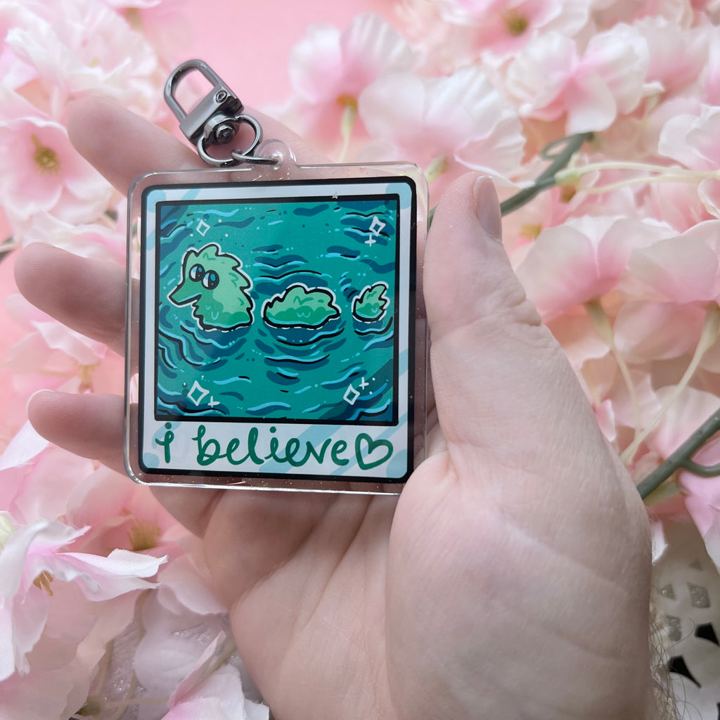 A keychain that looks like a camera photo, hand written text at the bottom that says "i believe <3", in the photo is lake water with a fuzzy worm lochness monster