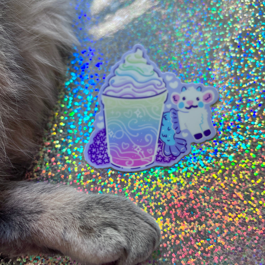 A colorful sticker of a rainbow-themed drink and mermaid goat on a glittery surface with a paw in the foreground.