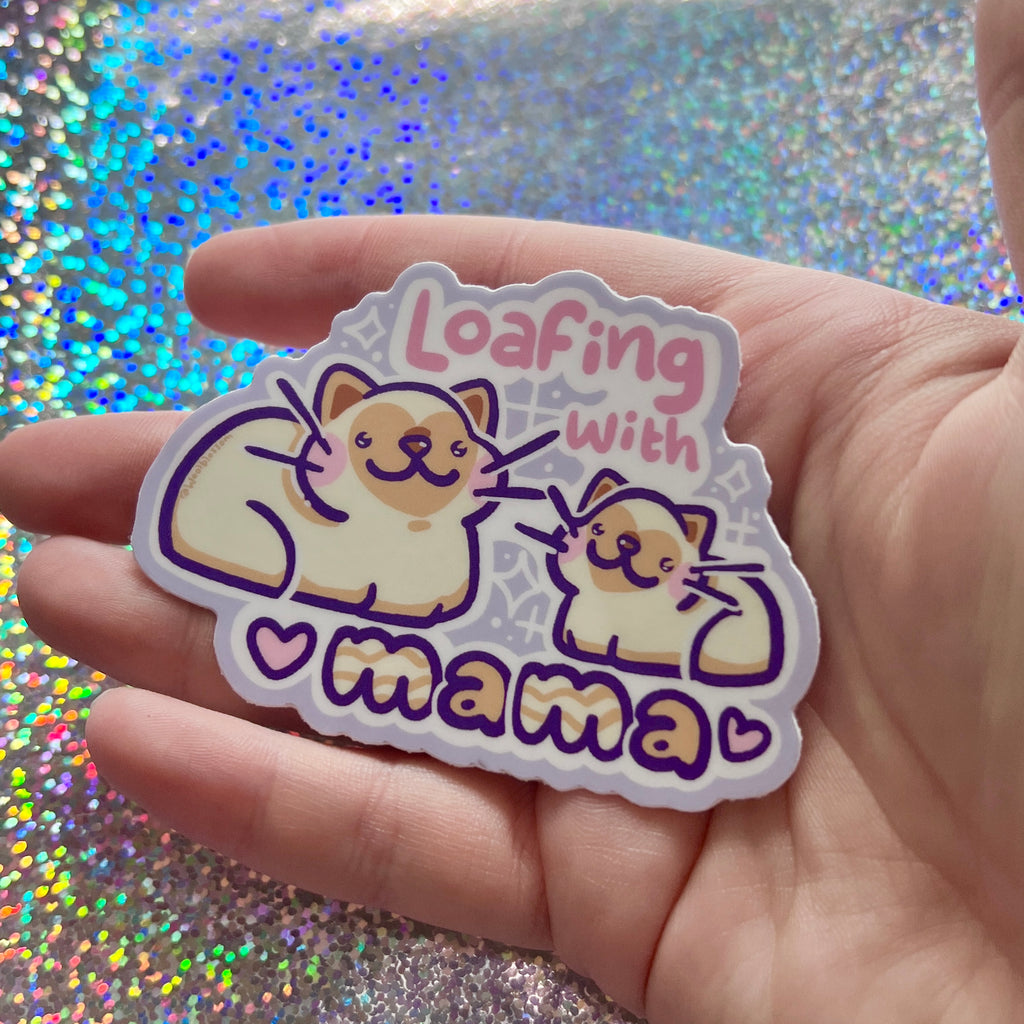 Sticker with cartoon dogs and text 'Loafing with mama' held in a hand against a glittery background.