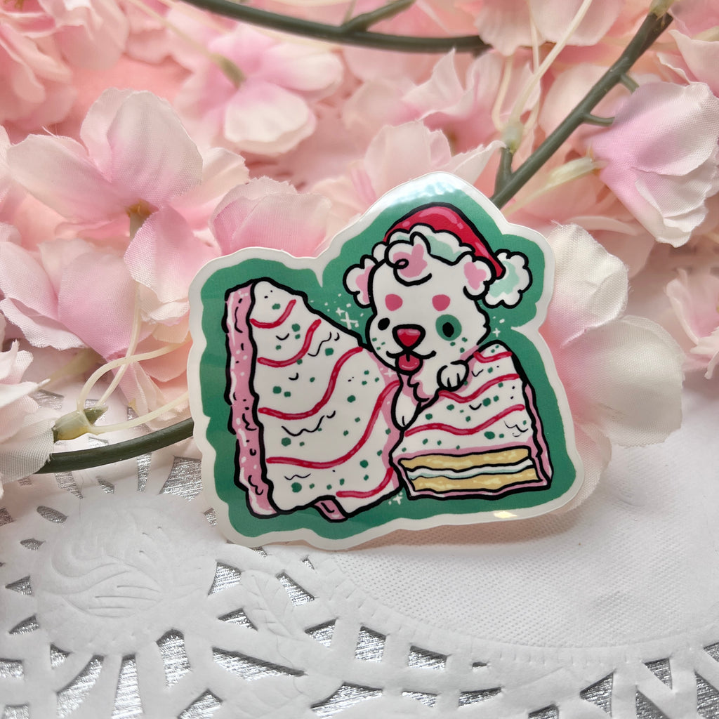 A sticker of a white puppy with a santa hat on top of two white christmas tree snack cakes