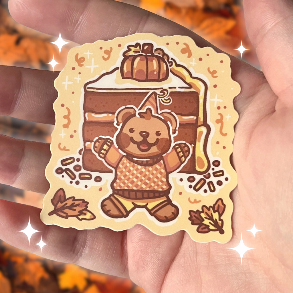 Sticker of a bear in an orange outfit with a pumpkin on top of a cake, held in a hand.