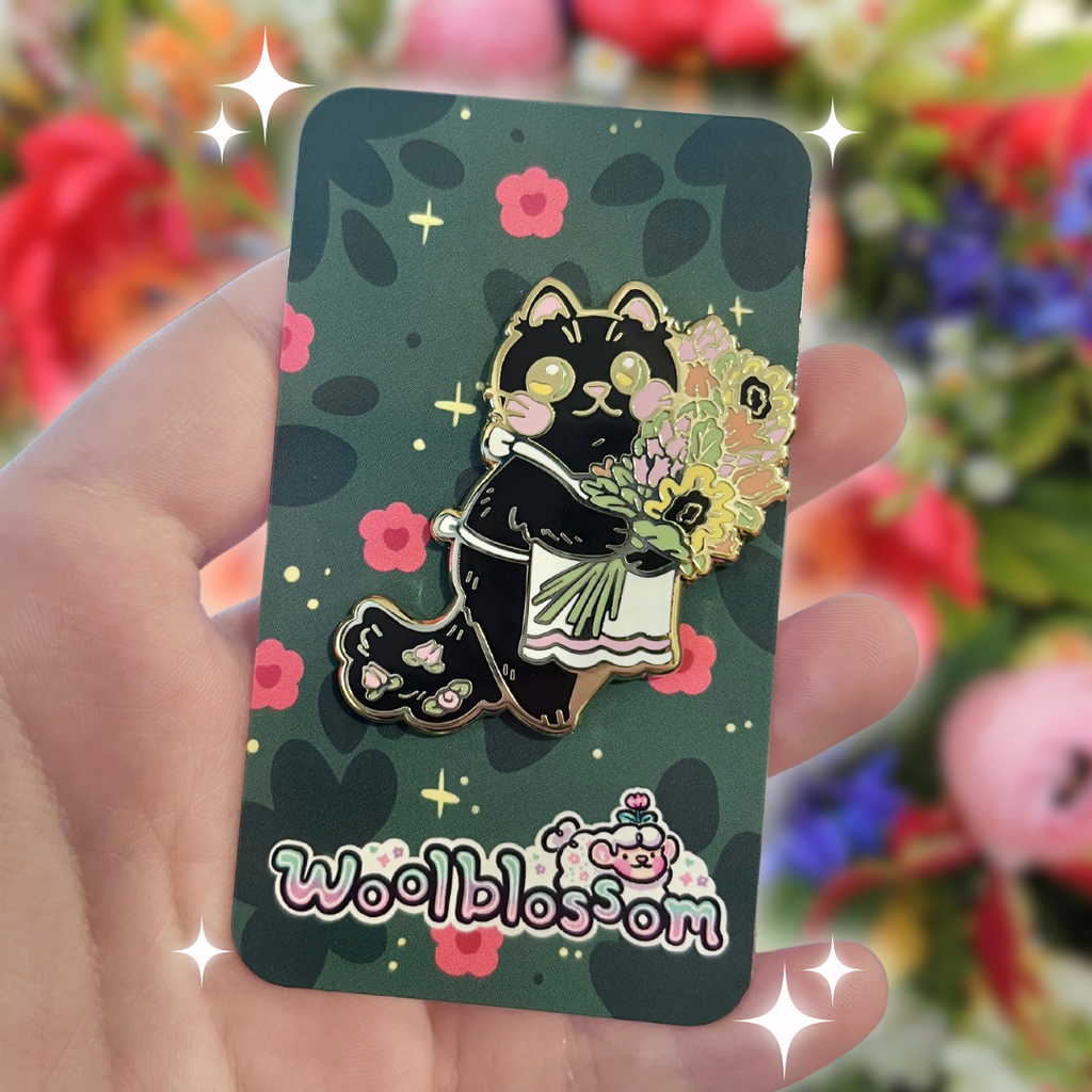 A gold enamel pin of a black cat, with pink details in his ears/nose/blush/little pink flowers in his tail, with a white apron, holding a bouquet of yellow/pink/orange flowers