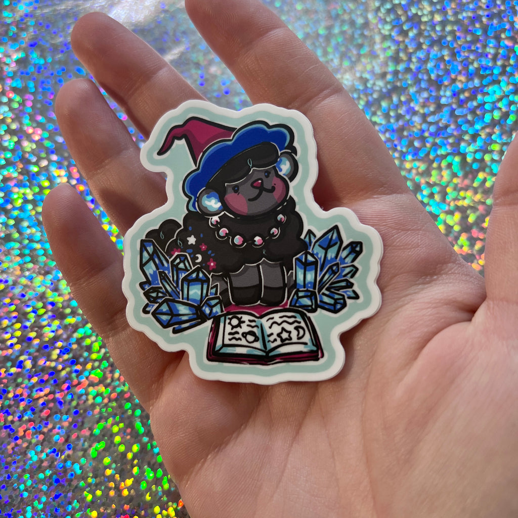 Hand holding a sticker of a sheep with crystals and a book against a glittery background