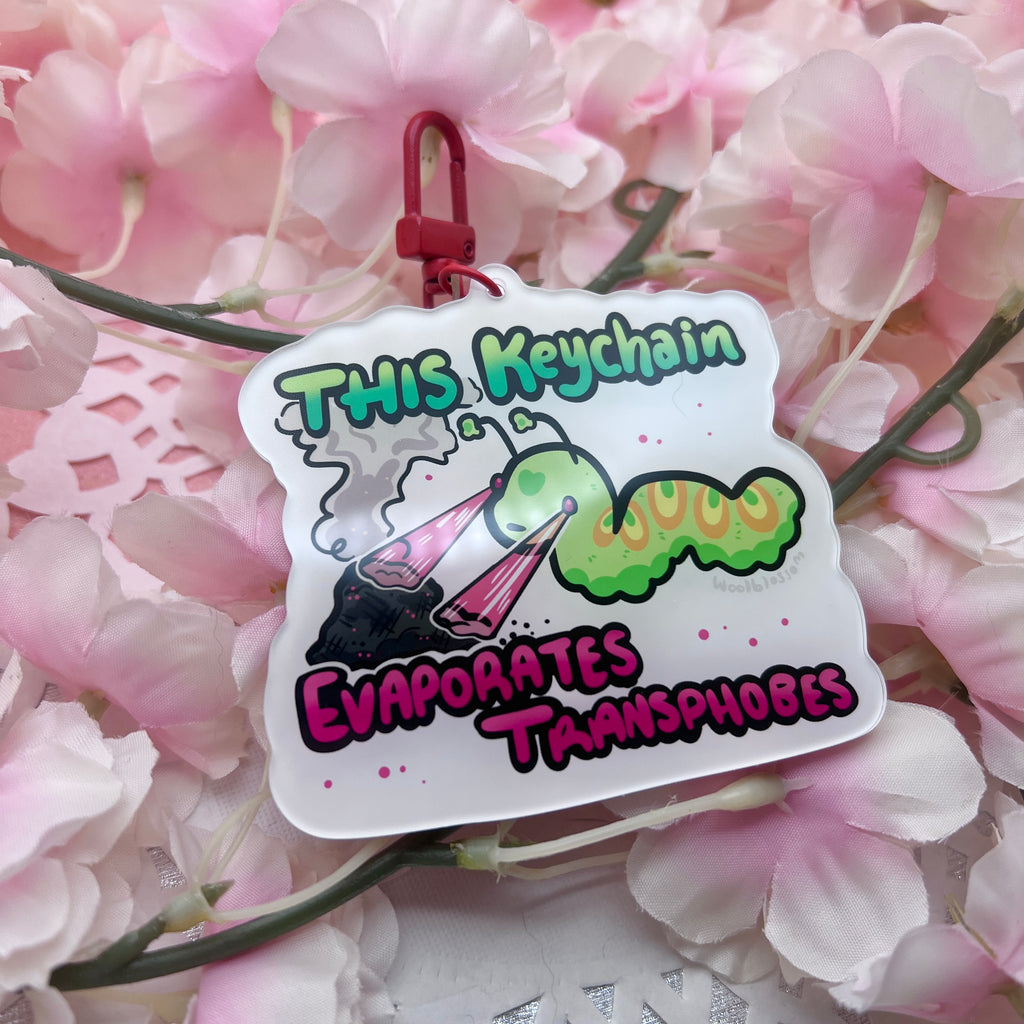 A keychain of a green worm shooting lasers out of its eyes onto a pile of ash with text that says "this keychain evaporates transphobes"