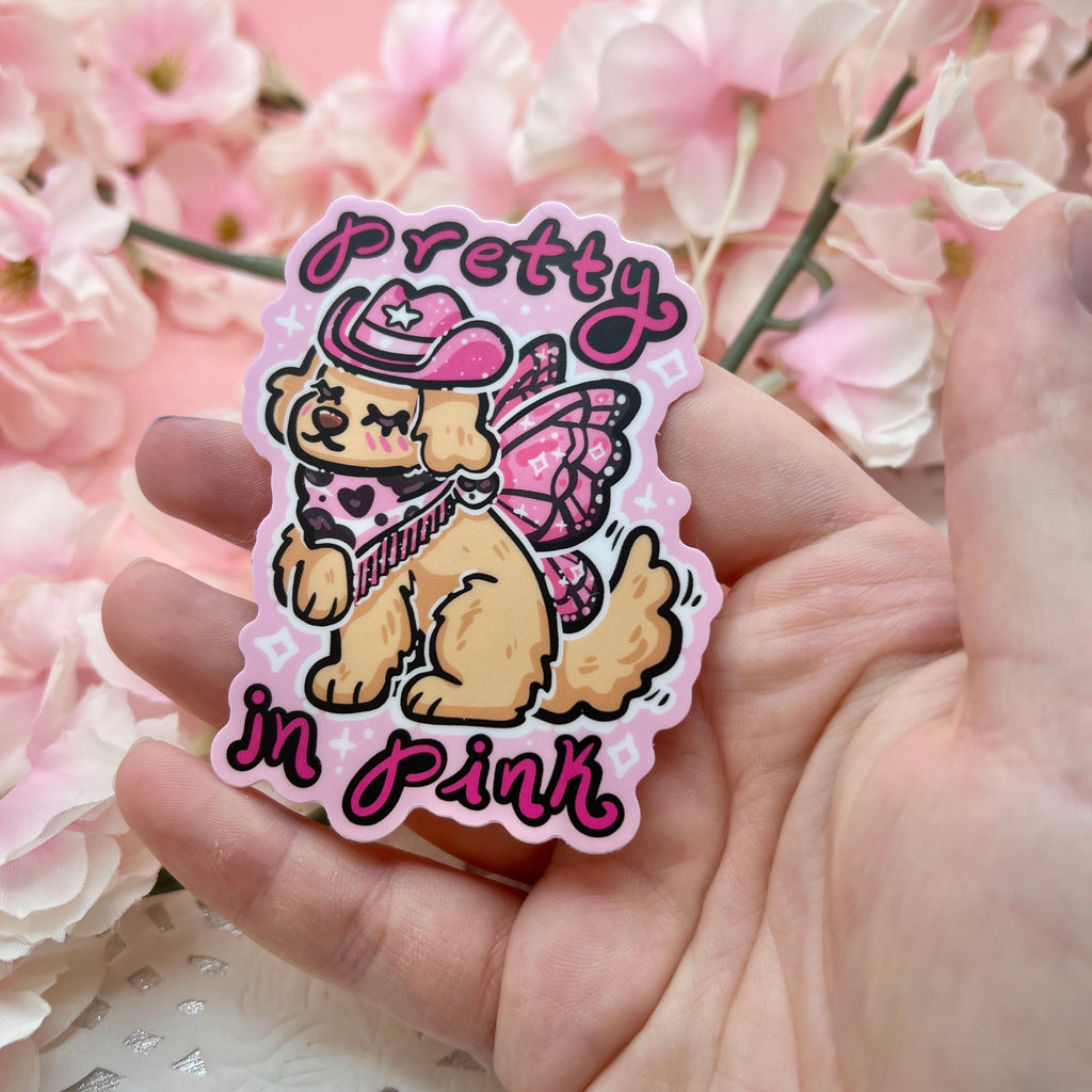 a sticker of a golden retreiver with a pink cowgirl hat, boots, and dark pink fairy wings and text that says "pretty in pink"