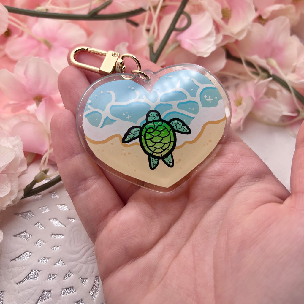 A keychain charm that is heart shaped, showing the ocean tide on sand, with a turtle in the middle