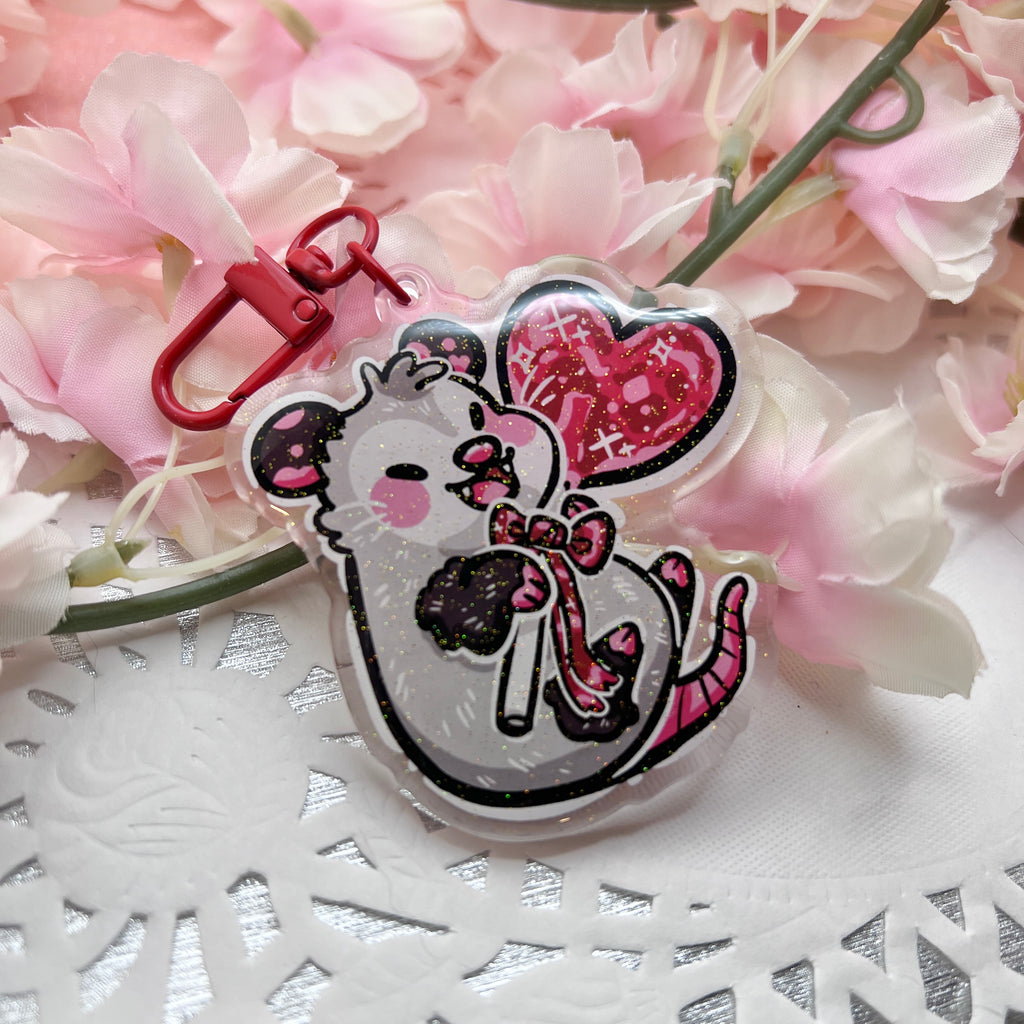 a keychain of an opossum happily holding a red heart lollipop