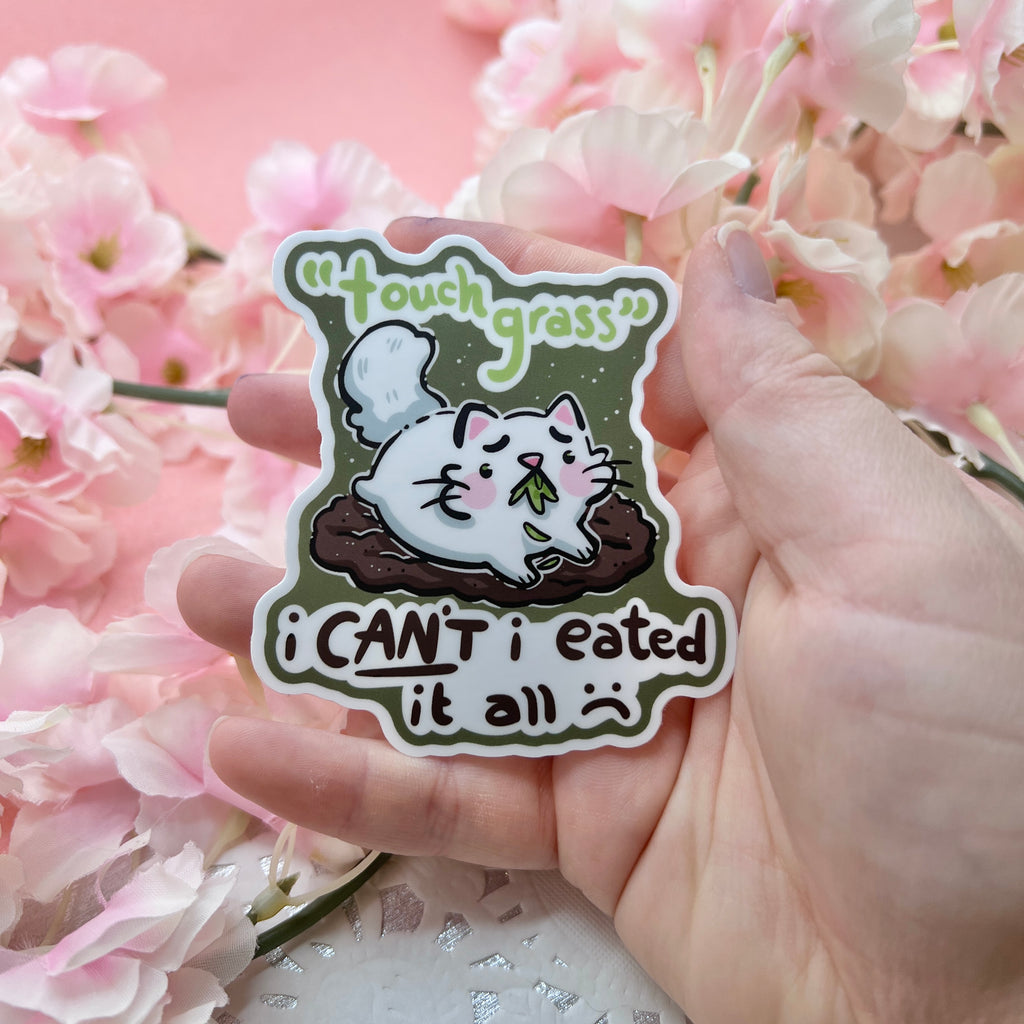 A sticker of a distressed cat laying on a bare patch of dirt, grass in her mouth with text that says "touch grass, i can't i eated it all :("