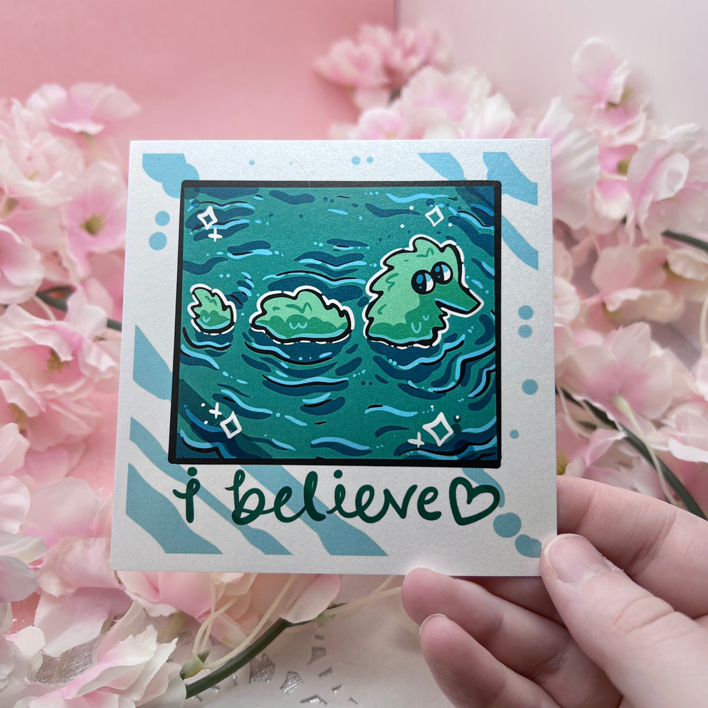 A print that looks like a camera photo, hand written text at the bottom that says "i believe <3", in the photo is lake water with a fuzzy worm lochness monster