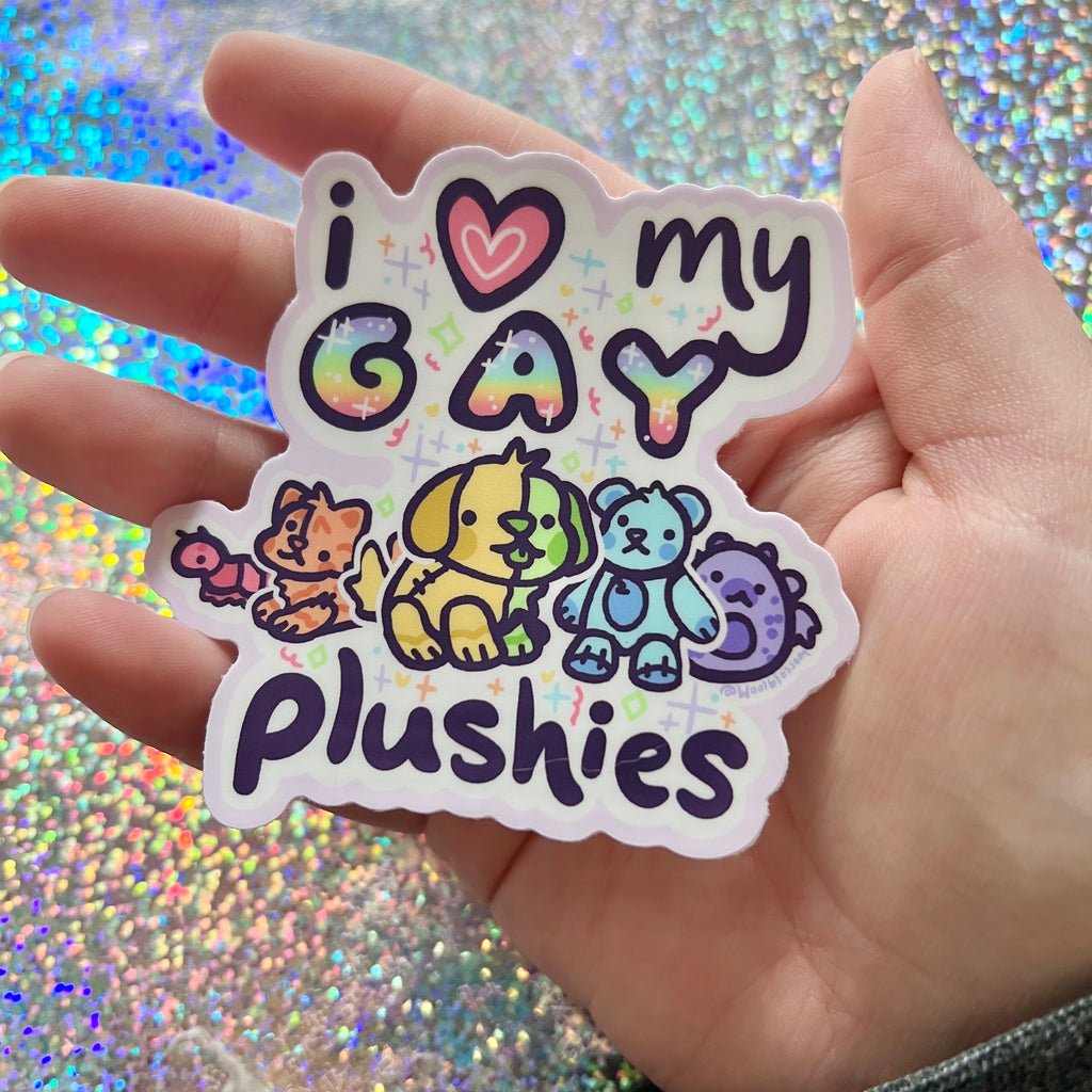 Hand holding a Sticker with 'i ❤️ my GAY plushies' text and rainbow-colored plushies on a glittery background against a glittery background