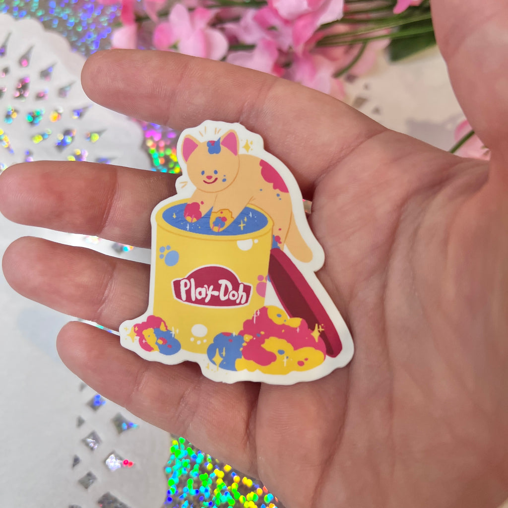 A hand is holding a Nostalgic Clay Cat ~ Sticker (v.3) featuring a professionally made illustration of a cute cartoon cat sitting inside a yellow Play-Doh tub, splattered with colorful paint. The sticker, printed on matte vinyl and water-proof, is displayed against a sparkly, holographic background with blurred pink flowers.