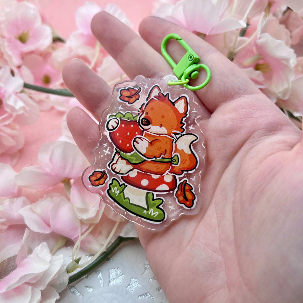 A keychain of a red fox with a little green apron sitting on a red mushroom, holding a strawberry with autumn leaves falling