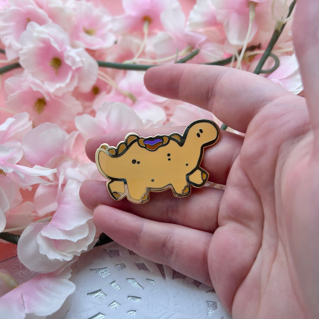 A gold enamel pin of a tan colored stegosaurus inspired dino that resembles a PBJ sandwich, the crustless kind, with a bite taken out of its back so you see the PB and jelly