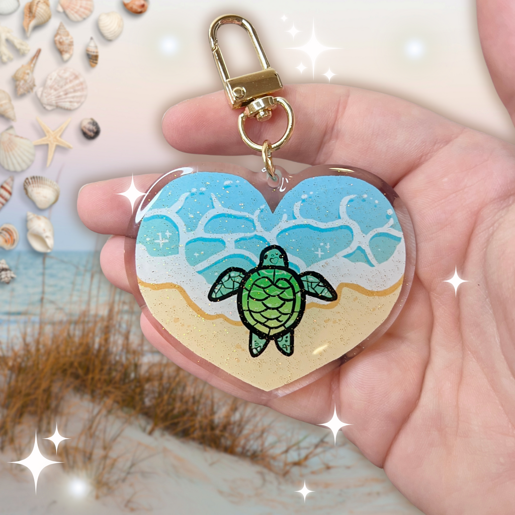 A keychain charm that is heart shaped, showing the ocean tide on sand, with a turtle in the middle