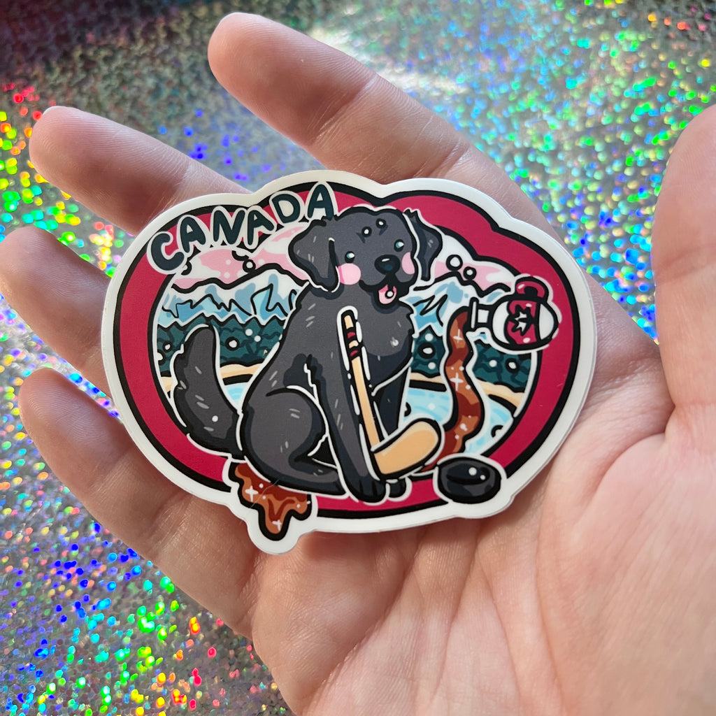 Hand holding a sticker with a black dog, hockey stick, and 'Canada' text against a glittery background.