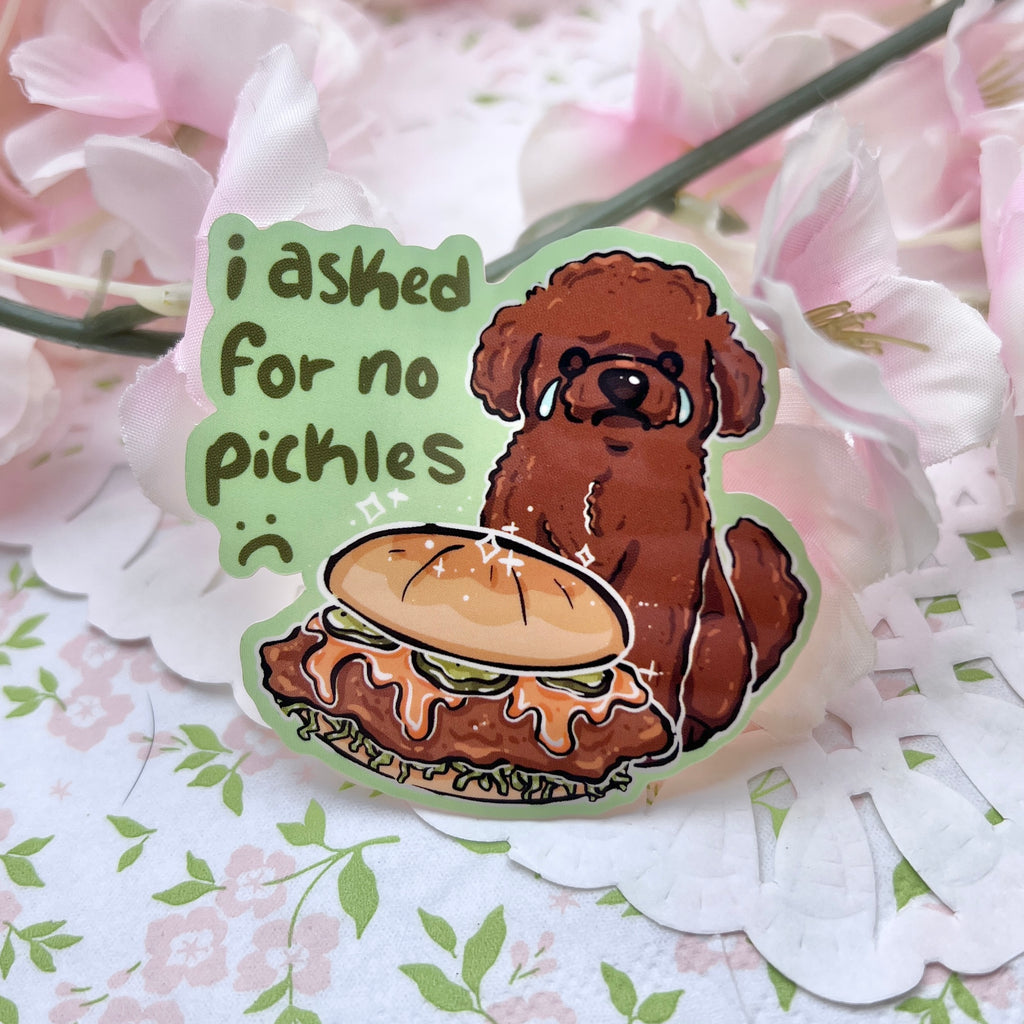 A sticker of a brown poodle dog sad and crying in front of a chicken sandwich that has pickles. Text says "i asked for no pickles :(" against a green background