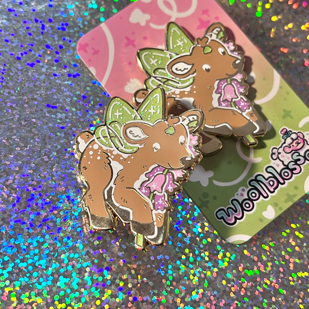 An enamel pin of a fairy deer holding a heather flower on a glittery surface with a colorful card in the background.