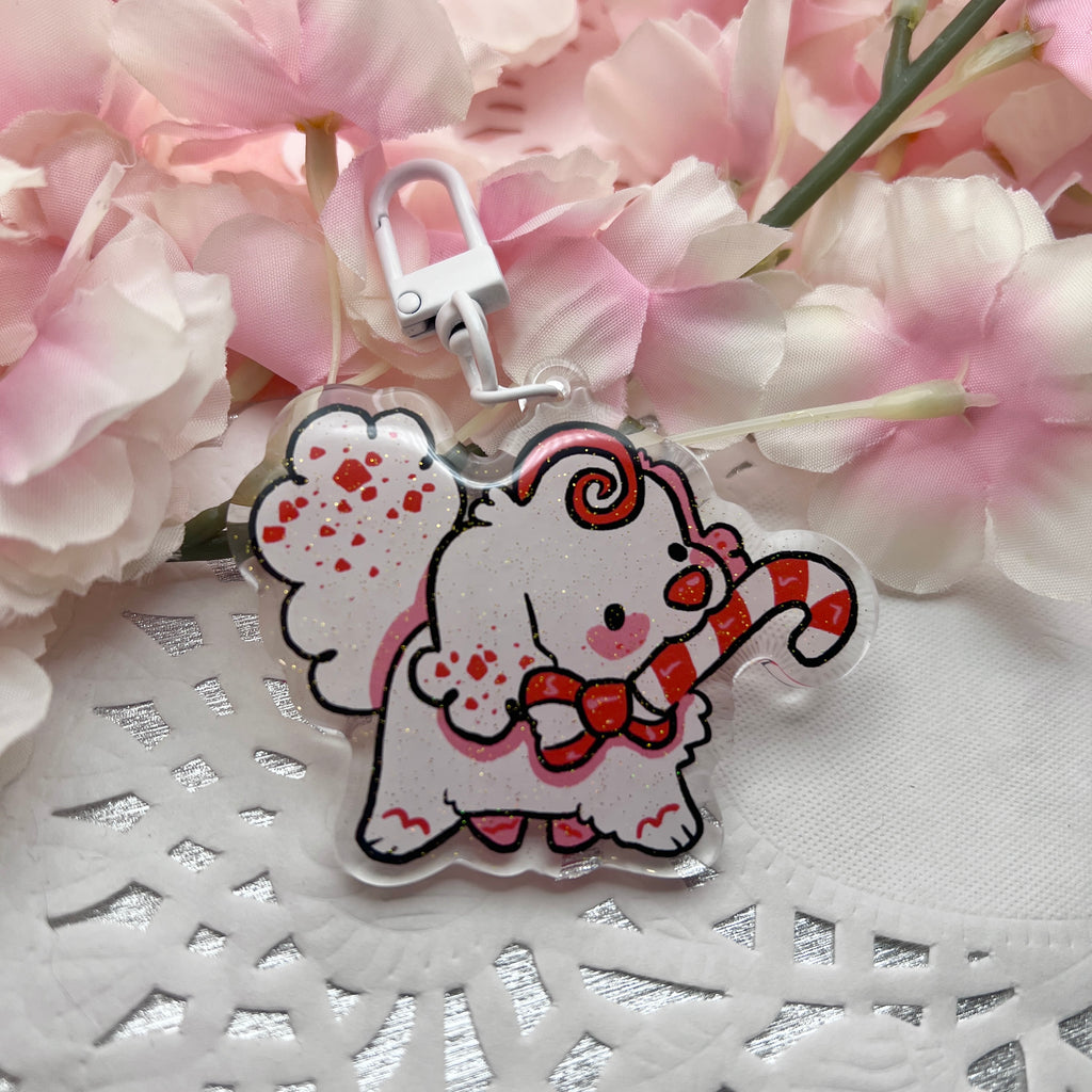 A keychain charm of a white peppermint bark themed puppy holding a candy cane in her mouth